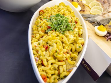 Weiße Schale mit gelbem Nudelsalat, garniert mit frischen Kräutern, auf dunklem Tuch neben Holzbrett mit Eierscheiben.