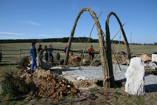 Mehrere Personen pflanzen einen Weidenpavillon mit gebogenen Weidenruten auf einem offenen Feld unter blauem Himmel