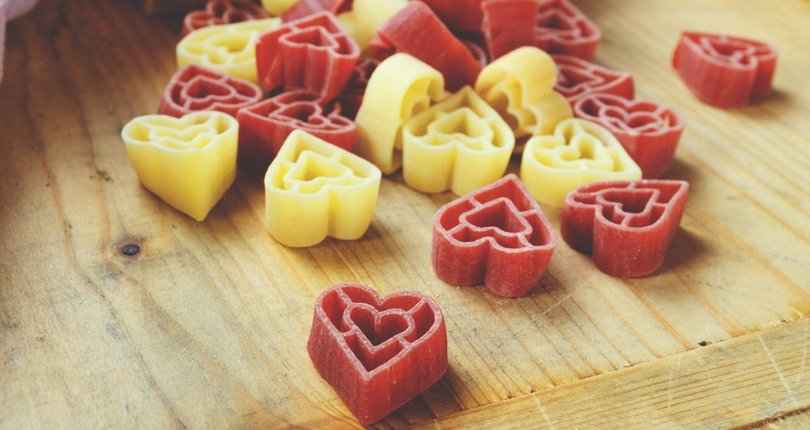Herzförmige Nudeln in Rot und Gelb, die aus einer geöffneten Verpackung auf einen Holztisch fallen.
