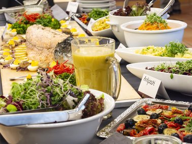 Buffet mit verschiedenen Salaten, Gemüse, einem Glas grüner Saft und mehreren weißen Schalen und Tellern auf einem Tisch.