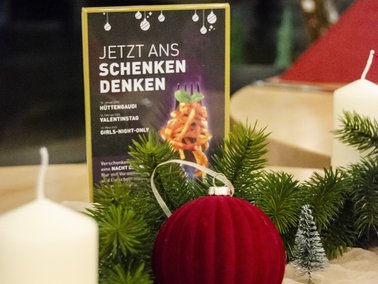 Rote Samtkugel, weiße Kerzen und Tannengrün vor einem schwarzen Schild mit weißer Aufschrift 'JETZT ANS SCHENKEN DENKEN'.