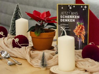 Roter Weihnachtsstern in braunem Blumentopf, weiße Kerze, kleine Tannenbäume und rote Weihnachtskugeln auf Tisch mit beige Stoffdecke.