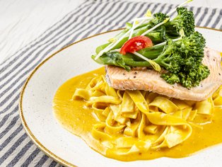 Gebratener Fisch auf Bandnudeln mit gelber Sauce, garniert mit Brokkoli, Salat und einer halben Kirschtomate auf weißem Teller.