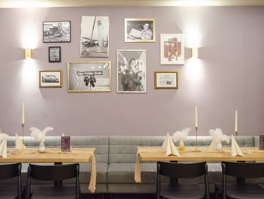 Wand mit mehreren gerahmten Schwarz-weiß-Fotografien über einer grauen Sitzbank und zwei Holztischen mit schwarzen Stühlen.