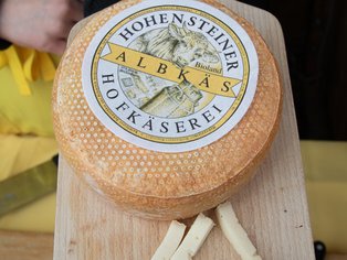 Runder Laib Käse mit gelber Rinde und Etikett 'Hohensteiner Albkas Hofkäserei' auf Holzbrett, davor einige Käsestücke.
