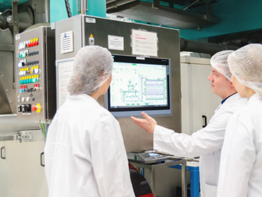 Drei Personen in weißen Schutzkitteln und Haarnetzen stehen vor einem großen Monitor mit technischen Diagrammen in einer Nudelproduktionsanlage.