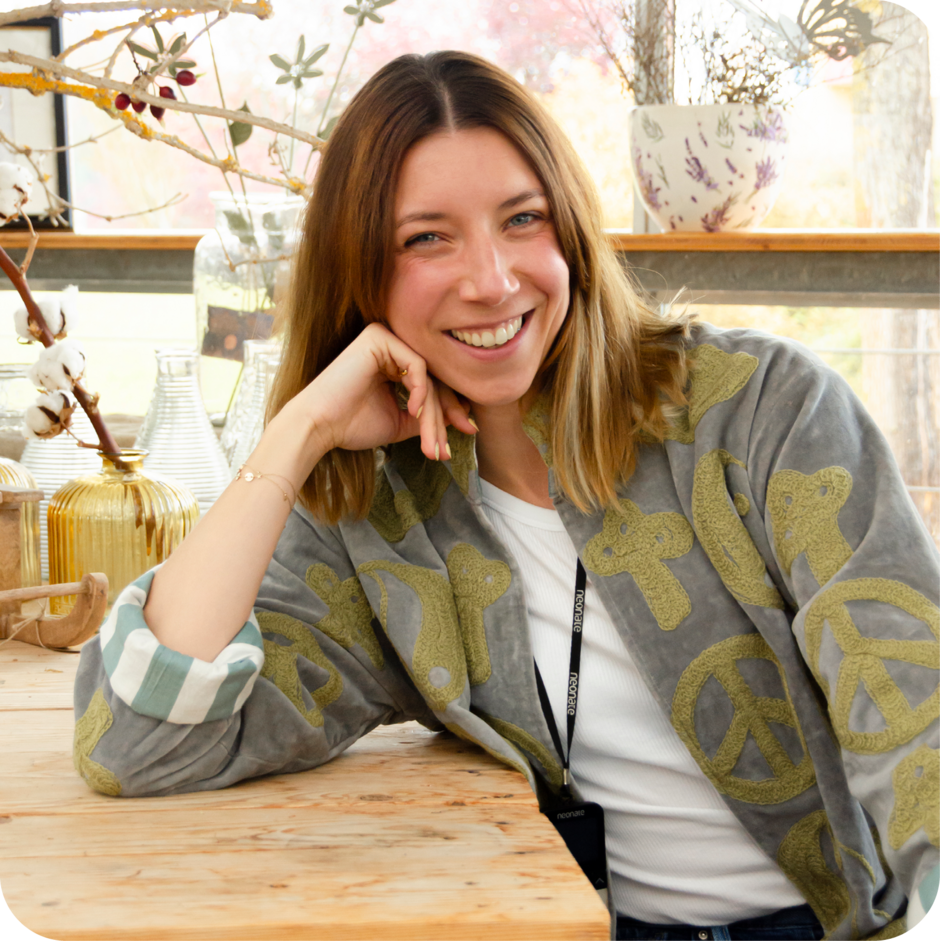 Person mit schulterlangem Haar lehnt mit dem Ellenbogen auf einem Holztisch, trägt graue Jacke mit Peace-Zeichen und weißes T-Shirt, im Hintergrund Pflanzen und Fenster.
