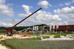Baustelle eines Pavillons mit rotem Kran, umgeben von grüner Wiese und Gebäuden im Hintergrund unter bewölktem Himmel