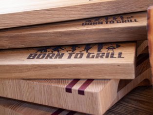 Mehrere übereinanderliegende Holzschneidebretter, eines mit der Aufschrift 'BORN TO GRILL' und Sternen in Großbuchstaben.