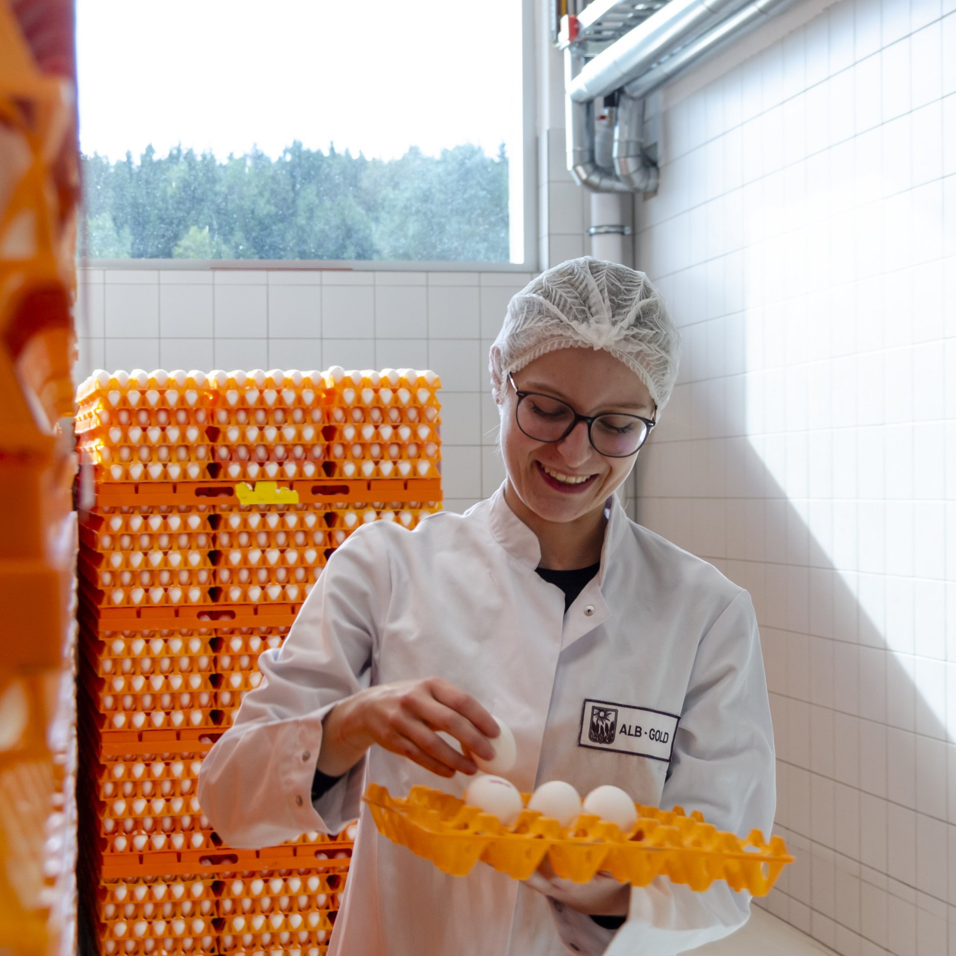 Person in weißem Kittel und Haarnetz hält ein Tablett mit mehreren weißen Eiern in einem Raum mit gestapelten orangen Eierbehältern.