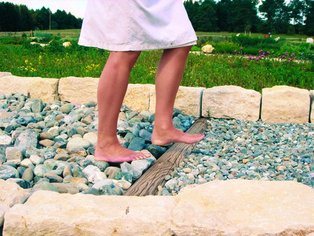 Beine einer Person in weißem Kleid, barfuß auf einem Kiesweg mit großen Steinen und Sand, im Hintergrund Wiese und Bäume.