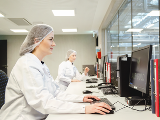 Zwei Personen in weißen Laborkitteln und Haarnetzen sitzen an Schreibtischen vor Computermonitoren in einem hellen Laborraum.
