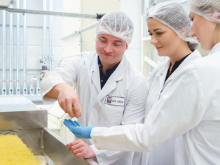 Zwei Personen in weißen Laborkitteln und Haarnetzen stehen an einer Produktionslinie mit gelbem Lebensmittel.