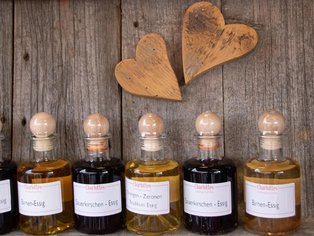 Fünf Flaschen mit Holzstopfen, gefüllt mit Flüssigkeiten in verschiedenen Gelb- und Brauntönen, vor einer Holzplatte mit zwei hölzernen Herzformen.