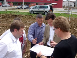 Vier Männer besprechen Pläne auf einer Baustelle mit gegrabenem Boden und parkenden Autos im Hintergrund Vier Männer besprechen Pläne auf einer Baustelle mit gegrabenem Boden und parkenden Autos im Hintergrund