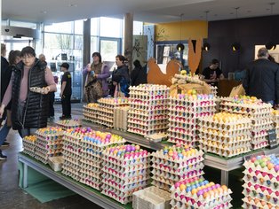 ehrere Tische mit großen Mengen bunt gefärbter Eier in Eierkartons in einem hellen Innenraum mit mehreren Personen im Hintergrund