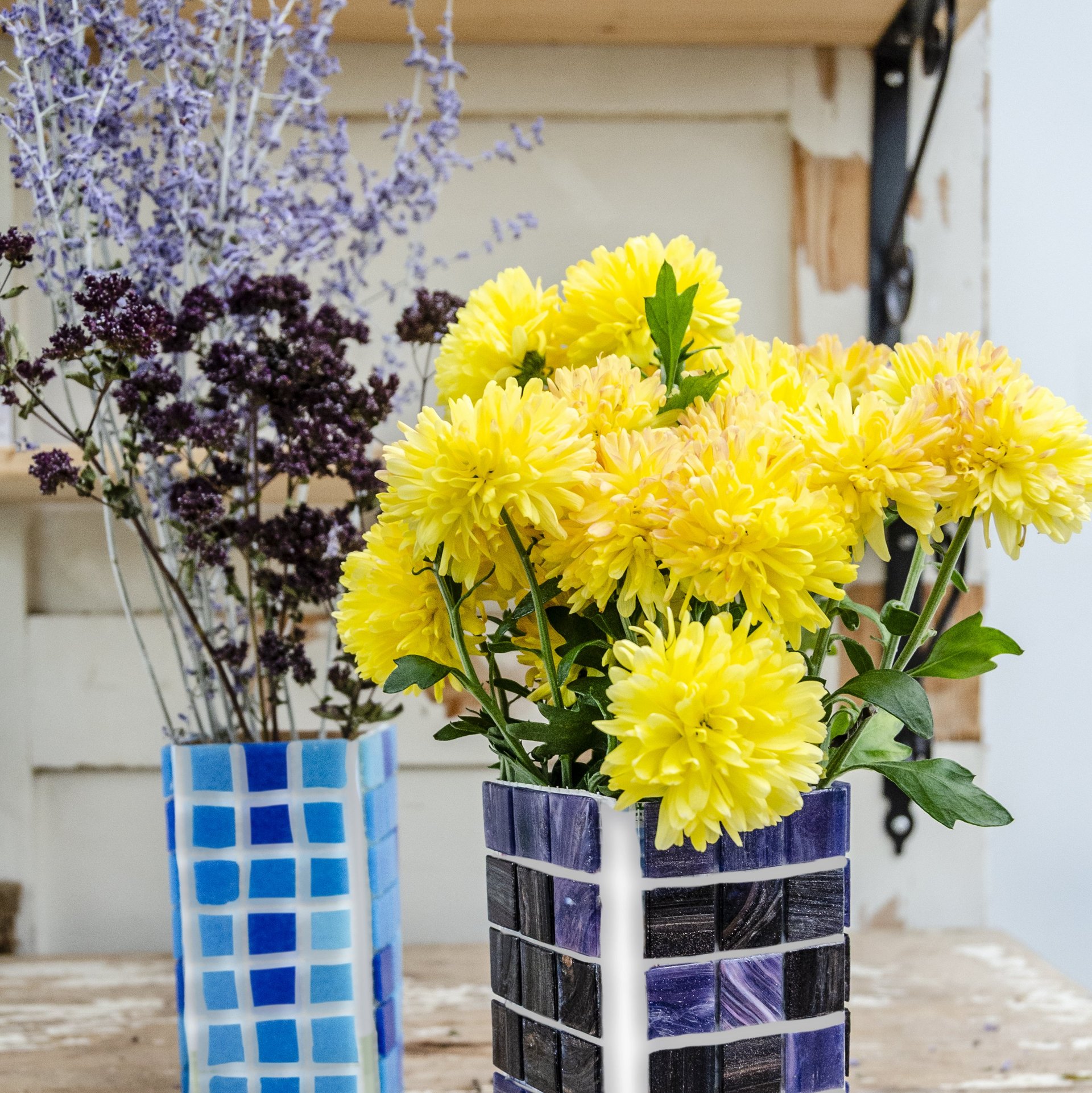 Gelbe Chrysanthemen in blau-schwarzer quadratischer Vase neben lila und dunkelroten Blüten in blau-weißer Vase auf Holztisch