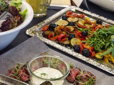 Vorspeisenteller mit dünn geschnittenem Rindfleisch, Sprossen und einem Glas mit weißer Sauce, daneben ein Tablett mit gegrilltem Gemüse und ein Salat in einer weißen Schüssel.