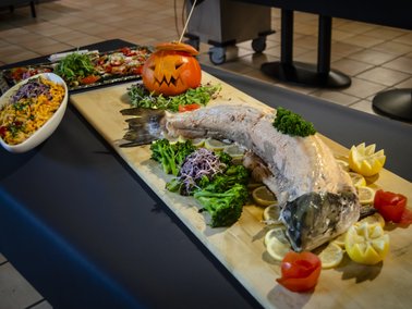 Langer Fisch auf Holzbrett mit grünen Kräutern, Zitronenscheiben und Tomaten, daneben ein orangefarbener Kürbis mit geschnitztem Gesicht.