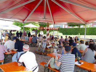 Menschen sitzen unter einem großen roten Sonnenschirm an orangenen Biertischgarnituren im Freien.