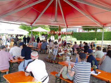 Menschen sitzen unter einem großen roten Pavillon auf Bänken an Tischen bei einer Veranstaltung mit kleinen Geschenken