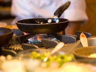 Zwei Pfannen auf einem Gasherd mit Flamme und unscharfem Koch im Hintergrund