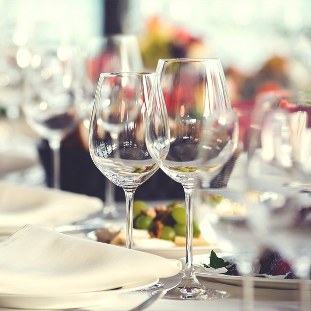 Weingläser auf gedecktem Tisch mit weißen Tellern und unscharfem Hintergrund mit Blumen und weiteren Gläsern