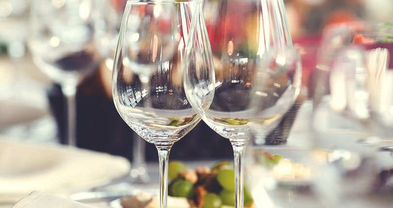 Weingläser auf gedecktem Tisch mit weißen Tellern und unscharfem Hintergrund mit Blumen und weiteren Gläsern