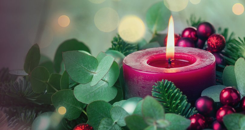 Brennende rote Kerze umgeben von Tannenzweigen, grünen Blättern und roten Weihnachtskugeln mit unscharfen Lichtern im Hintergrund.