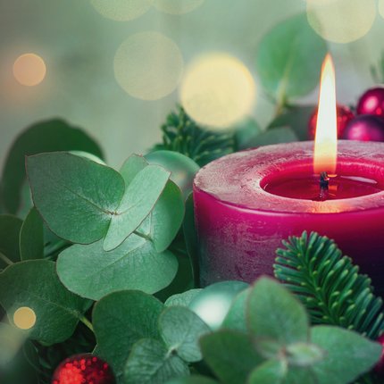Brennende rote Kerze umgeben von Tannenzweigen, grünen Blättern und roten Weihnachtskugeln mit unscharfen Lichtern im Hintergrund.