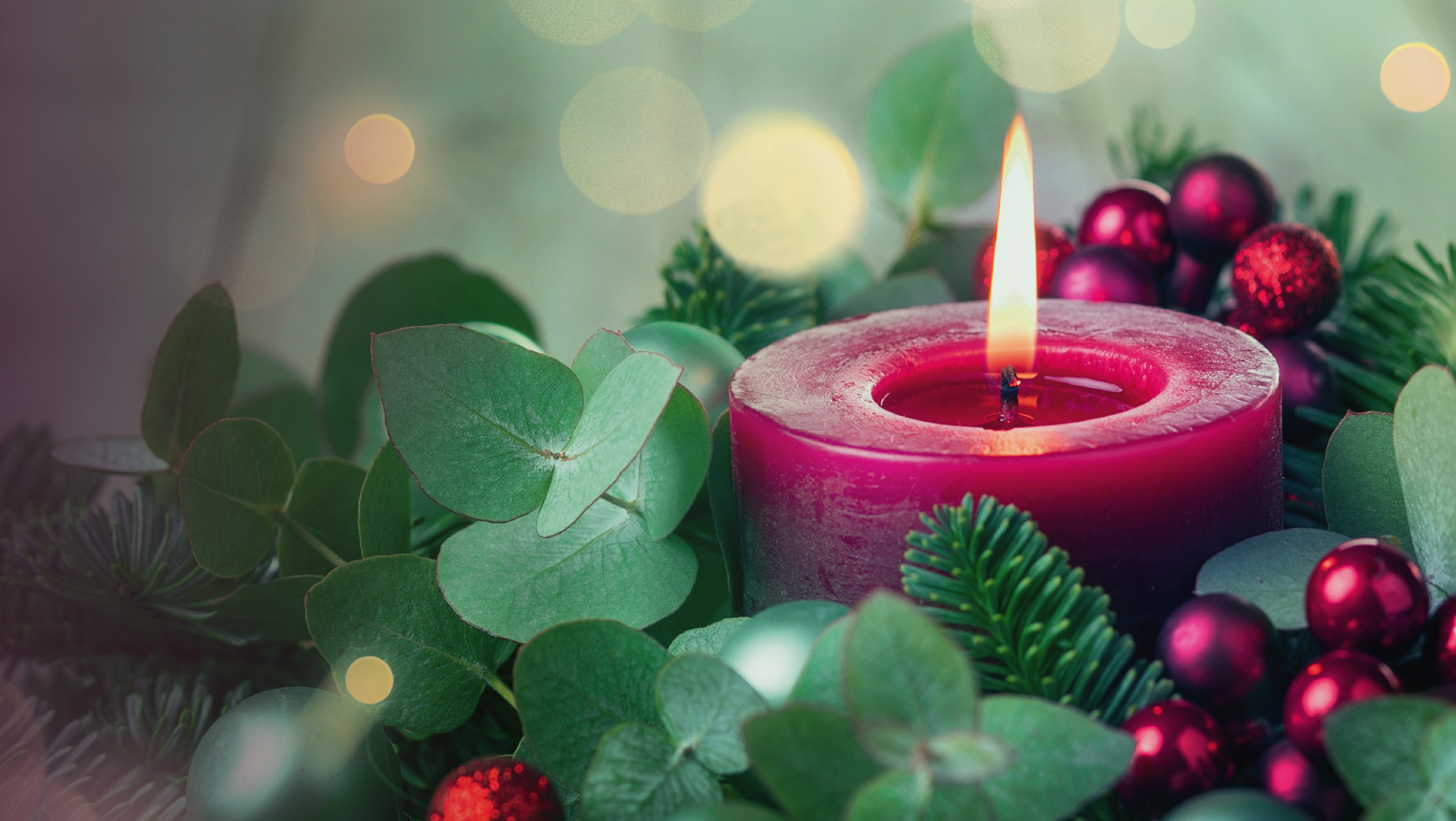 Brennende rote Kerze umgeben von Tannenzweigen, grünen Blättern und roten Weihnachtskugeln mit unscharfen Lichtern im Hintergrund.