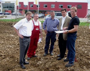 Fünf Männer stehen auf einer Baustelle, einer hält Pläne, im Hintergrund ein rotes Gebäude Fünf Männer stehen auf einer Baustelle, einer hält Pläne, im Hintergrund ein rotes Gebäude