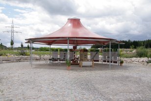 Runder Pavillon mit rotem Dach und offenen Seiten auf gepflastertem Platz unter bewölktem Himmel