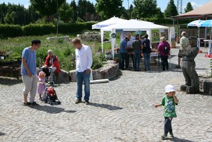 Menschen bei der Eröffnung eines Spielplatzes, darunter Erwachsene und Kinder, mit Pavillons und Skulpturen im Hintergrund
