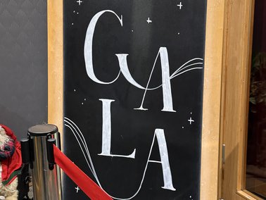 Schwarze Tafel mit weißer Schrift 'GALA' und dekorativen Linien, eingerahmt in Holz, neben rotem Absperrband.