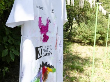 Ein weißes, selbst bemaltes T-Shirt hängt auf einer Schnur im Garten.