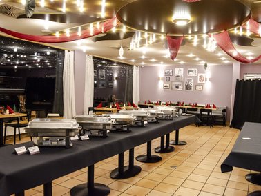 Buffet mit mehreren Warmhaltebehältern auf langen Tischen in einem festlich dekorierten Raum mit roten Girlanden und Lichterketten.