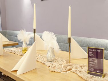 Drei gefaltete weiße Servietten, zwei hohe Kerzenhalter mit weißen Kerzen und zwei Vasen mit weißen Federn auf einem Holztisch.