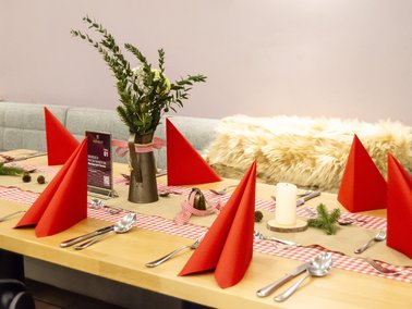 Tisch mit rot gefalteten Servietten, Besteck, Gläsern und einer Vase mit grünen Zweigen in einem hellen Raum.