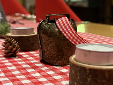 Auf rot-weiß kariertem Tischläufer steht eine braune Kuhglocke mit rotem kariertem Band, umgeben von Teelichtern in Holzhaltern und einer Tannenzapfen-Dekoration
