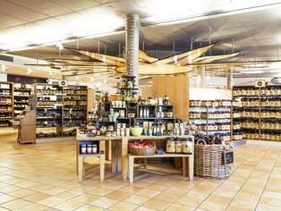Verkaufsraum mit Regalen und Tischen, auf denen verschiedene Lebensmittel und Produkte stehen, helle Decke mit sichtbaren Rohren