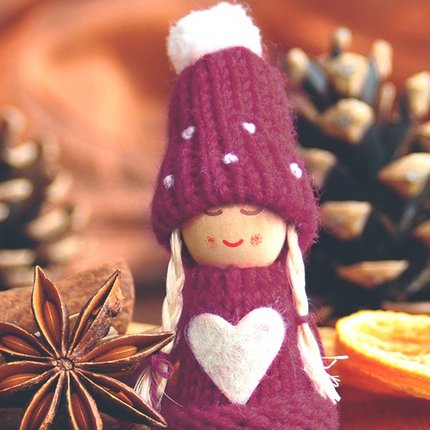 Figur mit lila Mütze und Pullover mit weißem Herz, umgeben von Zimtstangen, Sternanis, Kaffeebohnen, getrockneter Orangenscheibe und Kiefernzapfen auf Holzuntergrund.