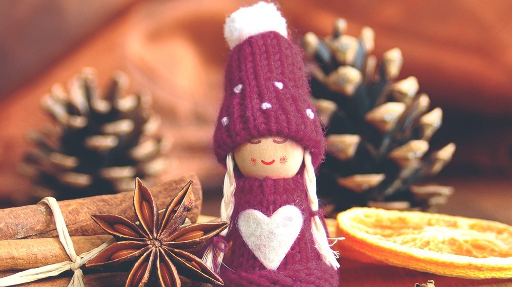 Figur mit lila Mütze und Pullover mit weißem Herz, umgeben von Zimtstangen, Sternanis, Kaffeebohnen, getrockneter Orangenscheibe und Kiefernzapfen auf Holzuntergrund.