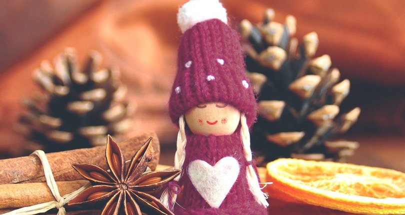 Figur mit lila Mütze und Pullover mit weißem Herz, umgeben von Zimtstangen, Sternanis, Kaffeebohnen, getrockneter Orangenscheibe und Kiefernzapfen auf Holzuntergrund.