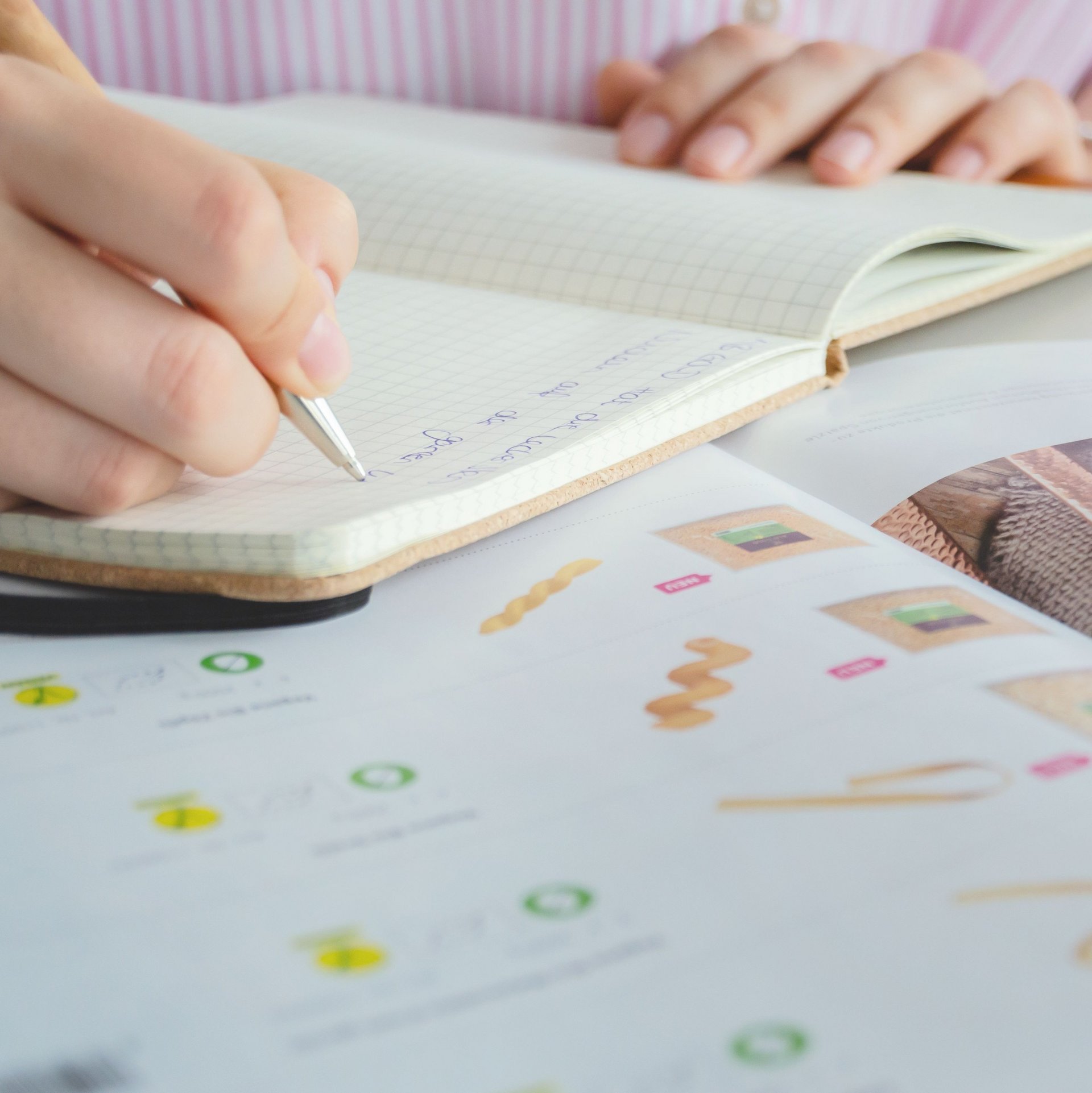 Person schreibt mit Stift in ein Notizbuch, daneben liegt ein aufgeschlagenes Magazin mit Bildern und Text