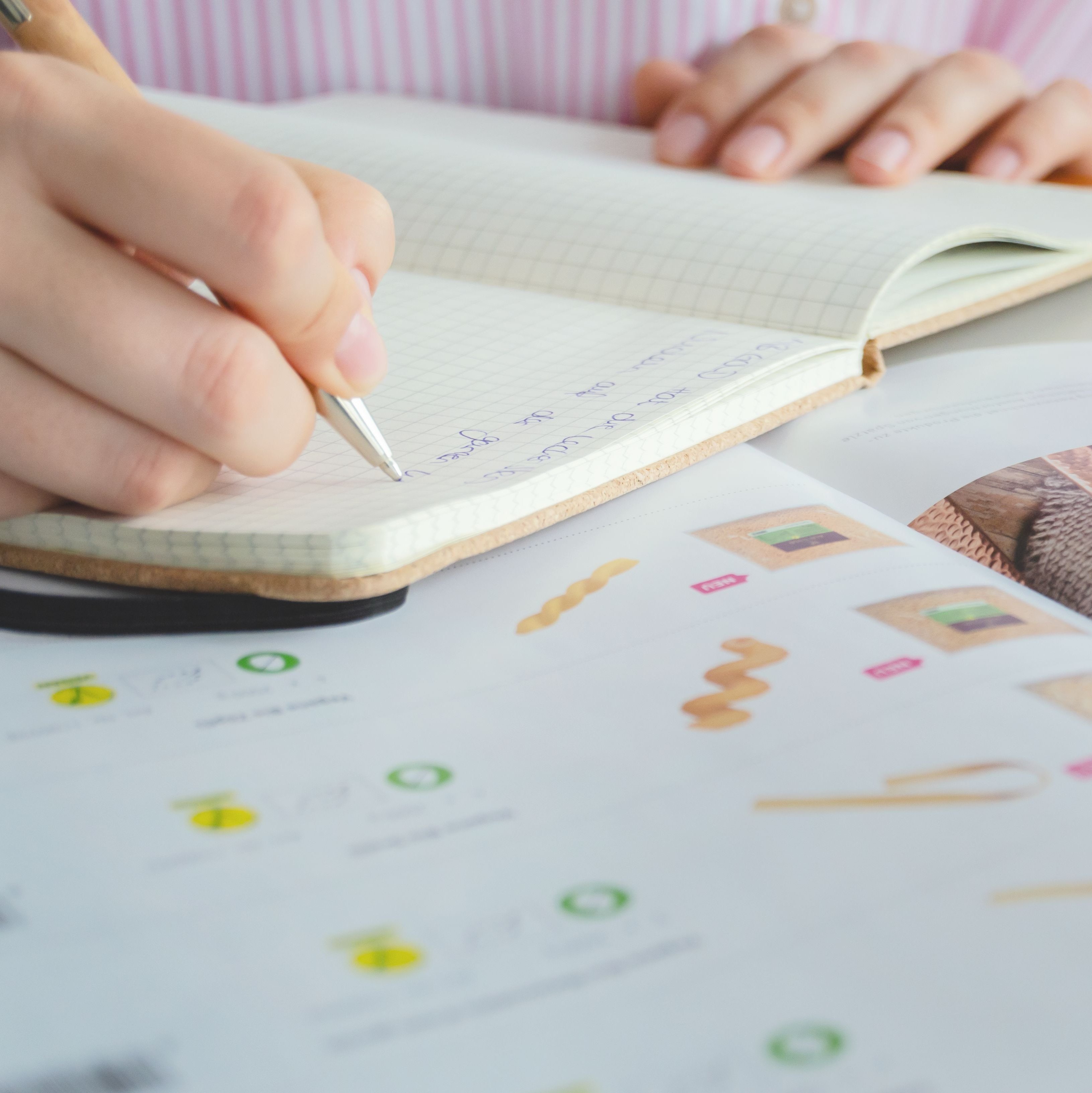 Person schreibt mit Stift in ein Notizbuch, daneben liegt ein aufgeschlagenes Magazin mit Bildern und Text