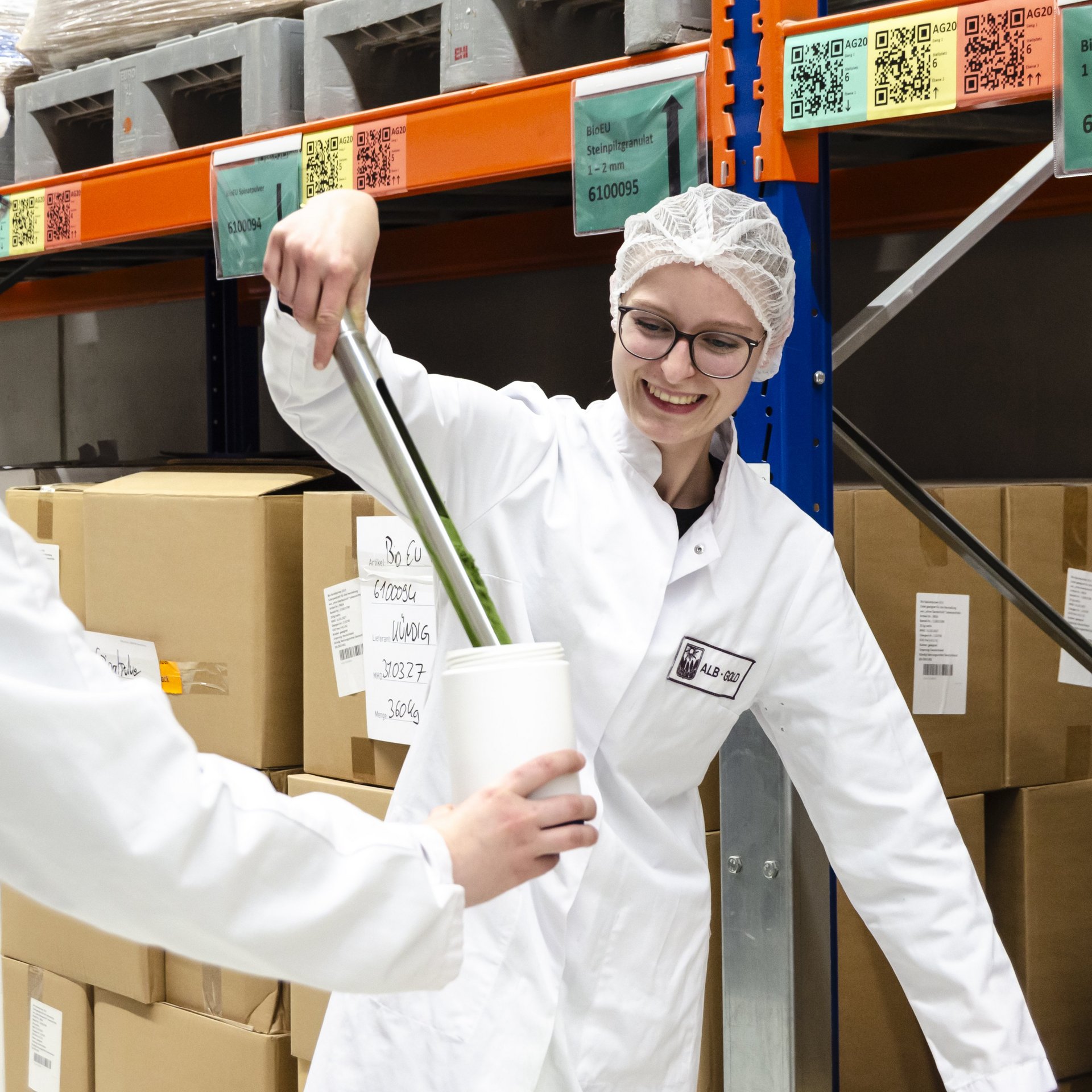 Zwei Personen in weißen Laborkitteln und Haarnetzen in einem Lager, eine hält einen Becher, die andere benutzt ein Rohr welches in das Gefäß ragt..