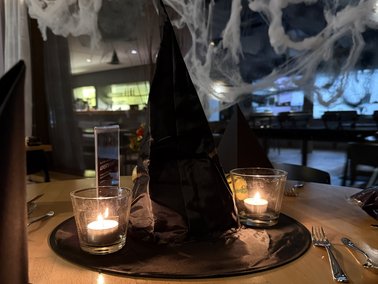 Dunkler, spitzer Hexenhut auf einem runden Teller, umgeben von zwei brennenden Teelichtern auf einem gedeckten Tisch.