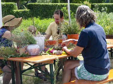 Erwachsene Personen sitzen auf einer Bierbank und basteln Blumenkränze