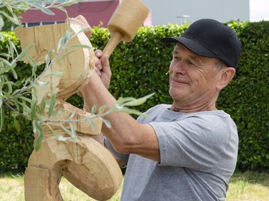 Person mit grauem T-Shirt und schwarzer Mütze schnitzt eine Holzskulptur mit Hammer und Meißel im Garten